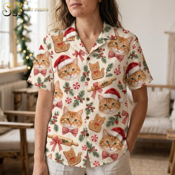 A woman wearing a hawaiian christmas shirt womens style, featuring large orange tabby cat faces, gingerbread cat cookies, rolling pins with red gingham ribbons, and holly sprigs, photographed from the waist up in a softly lit living room.
