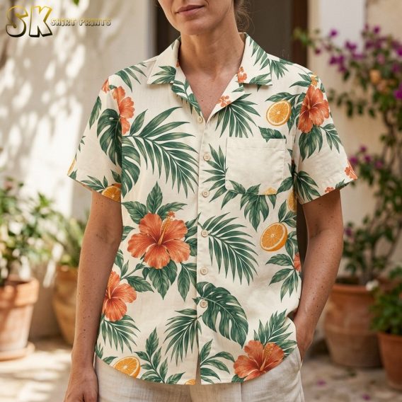 A woman wearing the cream tropical shirt stands in a sunlit courtyard, with the chest patch pocket and the orange hibiscus and fruit print clearly visible at close range.