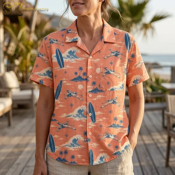 A woman wears a coral pink Hawaiian button-up shirt with a spacecraft-patterned chest pocket and blue surfboard illustrations.