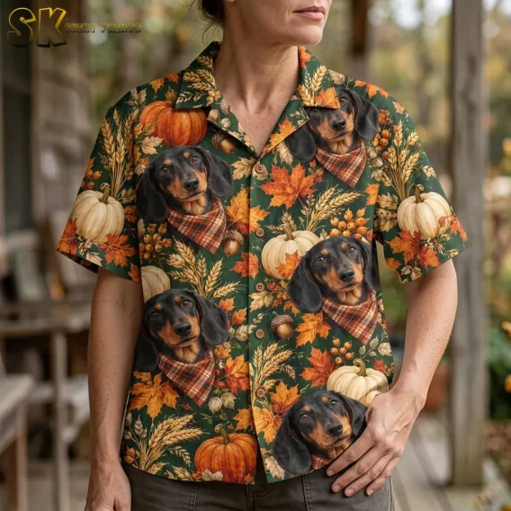 A woman wears a green button-up shirt with large dachshunds in plaid bandanas, white pumpkins, and orange leaves.