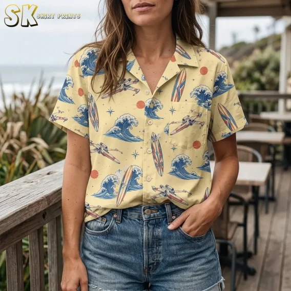 A woman wears the pale yellow printed button-up tucked into denim shorts, with blue waves and a spacecraft visible on the chest pocket.