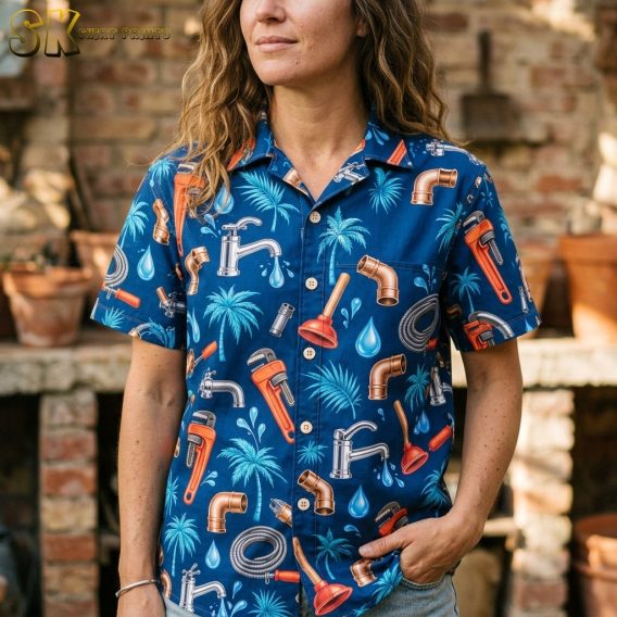 A woman with curly hair wearing a navy button-up shirt covered in plumbing motifs stands in front of a brick fireplace, with copper pipe elbows and red plungers clearly visible at close range.