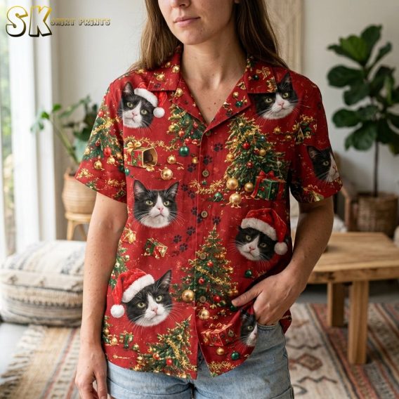 A woman wearing the red hawaiian christmas shirt indoors holds the hem, bringing the lower front panel into focus and highlighting the Santa-hat cat faces and Christmas tree details up close.