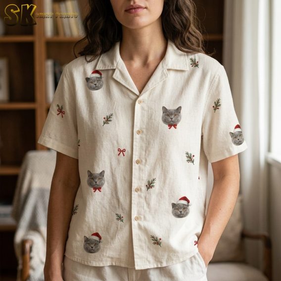 A woman wearing the cream womens hawaiian christmas shirt indoors, with the camera close enough to distinguish individual cat faces alternating between red bow ties and Santa hats, plus small holly sprigs between them.